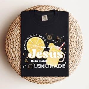 Puede incluir: Camiseta negra con un dise&ntilde;o gr&aacute;fico que presenta una rodaja de lim&oacute;n, un vaso de limonada con una pajita y el texto "Life gave me some lemons but my Jesus He be makin' lemonade."