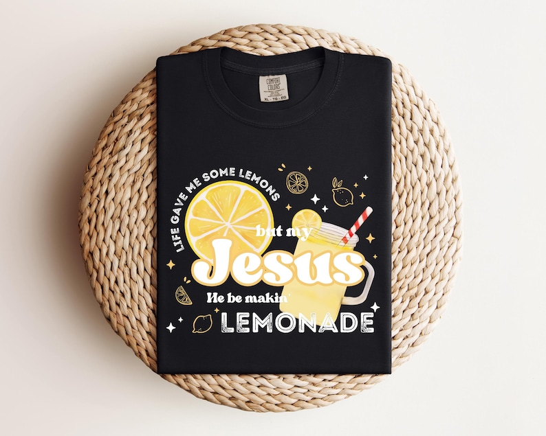 Puede incluir: Camiseta negra con un dise&ntilde;o gr&aacute;fico que presenta una rodaja de lim&oacute;n, un vaso de limonada con una pajita y el texto "Life gave me some lemons but my Jesus He be makin' lemonade."