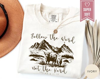 May include: Ivory-colored t-shirt with the text "Follow the word not the Herd" and a graphic of mountains and Highland cattle. The shirt is made of a soft material and has a crew neck.