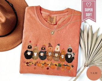 May include: A coral-colored Comfort Colors t-shirt featuring a whimsical design of chickens wearing hats and holding mugs of coffee. The graphic includes pumpkins and autumn leaves. The shirt has a round neckline and a gold necklace. The text "SUPER SOFT" is visible.