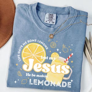May include: A blue Comfort Colors t-shirt with the text "Life gave me some lemons but my Jesus He be makin' lemonade." The graphic includes a large yellow lemon slice and a lemonade drink. A gold necklace is draped across the shirt.