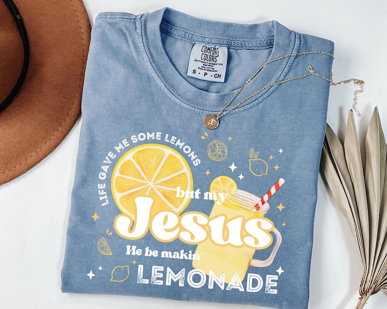 Puede incluir: Una camiseta azul Comfort Colors con el texto "Life gave me some lemons but my Jesus He be makin' lemonade." El gr&aacute;fico incluye una gran rodaja de lim&oacute;n amarillo y una bebida de limonada. Un collar de oro est&aacute; drapeado sobre la camisa.