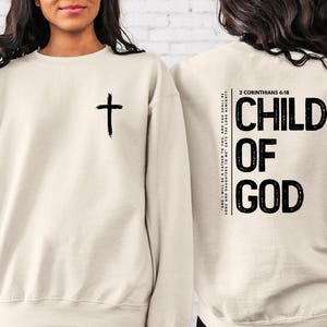 May include: A beige sweatshirt with a black cross on the front and the text "CHILD OF GOD" and the Bible verse "2 Corinthians 6:18" on the back.