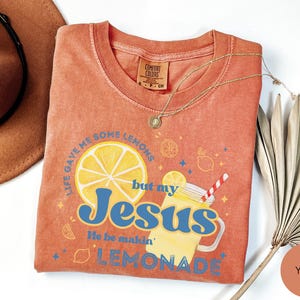 Puede incluir: Una camiseta de color coral con el texto "Life gave me some lemons but my Jesus He be makin' lemonade". El gr&aacute;fico incluye una rodaja de lim&oacute;n, un vaso de limonada y otras ilustraciones con tem&aacute;tica de lim&oacute;n. Un collar de oro est&aacute; drapeado sobre la camisa.