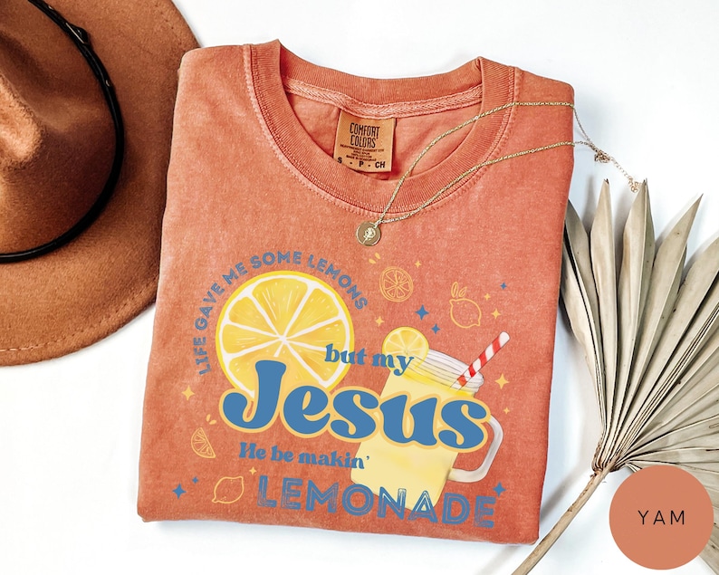 Puede incluir: Una camiseta de color coral con el texto "Life gave me some lemons but my Jesus He be makin' lemonade". El gr&aacute;fico incluye una rodaja de lim&oacute;n, un vaso de limonada y otras ilustraciones con tem&aacute;tica de lim&oacute;n. Un collar de oro est&aacute; drapeado sobre la camisa.