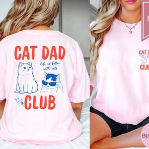 May include: A light pink t-shirt with the words "CAT DAD CLUB" in red. The back of the shirt features two cat illustrations and the phrase "life is better with cats." The front has a smaller "CAT DAD CLUB" design.