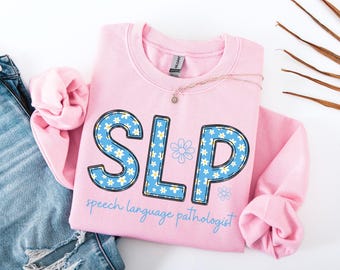 May include: A light pink sweatshirt with the letters "SLP" in a blue floral pattern. The words "speech language pathologist" are written in light blue script below the letters. A gold necklace with a small pendant rests on the sweatshirt.