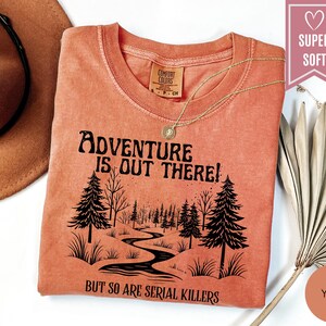 May include: A yam-colored t-shirt with the text "Adventure is Out There!" and "But So Are Serial Killers" in black. The graphic features a forest scene with a winding river. A gold necklace and a brown hat are also visible.
