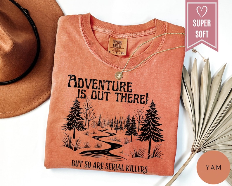 May include: A yam-colored t-shirt with the text "Adventure is Out There!" and "But So Are Serial Killers" in black. The graphic features a forest scene with a winding river. A gold necklace and a brown hat are also visible.