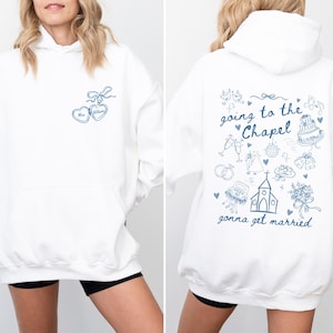 K&ouml;nnte beinhalten: Wei&szlig;er Kapuzenpullover mit blauen herzf&ouml;rmigen Anh&auml;ngern und dem Namen "Mrs. Wilson". Auf der R&uuml;ckseite des Hoodies steht der Text "going to the Chapel gonna get married" mit Illustrationen zum Thema Hochzeit. Ein l&auml;ssiges, bequemes Kleidungsst&uuml;ck.