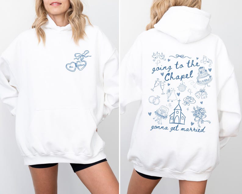 K&ouml;nnte beinhalten: Wei&szlig;er Kapuzenpullover mit blauen herzf&ouml;rmigen Anh&auml;ngern und dem Namen "Mrs. Wilson". Auf der R&uuml;ckseite des Hoodies steht der Text "going to the Chapel gonna get married" mit Illustrationen zum Thema Hochzeit. Ein l&auml;ssiges, bequemes Kleidungsst&uuml;ck.