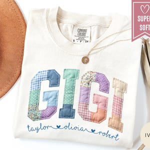 May include: Ivory-colored t-shirt with the name "GIGI" in patchwork letters. The letters are in various colors and patterns. Below the name are the names "taylor", "olivia", and "robert". A gold necklace is visible.