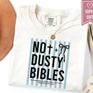 May include: An ivory t-shirt featuring a graphic with light blue vertical stripes and black text that reads NO DUSTY BIBLES, with a cross and a bow icon. Below the main text, smaller black text reads No Dusty Bible, No Empty Faith, Just A Heart Filled With God's Word.