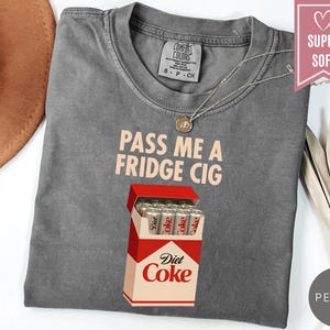 May include: A gray Comfort Colors t-shirt with the text "PASS ME A FRIDGE CIG" and a graphic of a Diet Coke cigarette pack. The shirt has a crew neck and short sleeves. A silver necklace is visible.