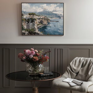 May include: A framed painting of a coastal town with buildings on a cliff overlooking a blue ocean. The painting is in a light wood frame and is hanging on a gray wall.