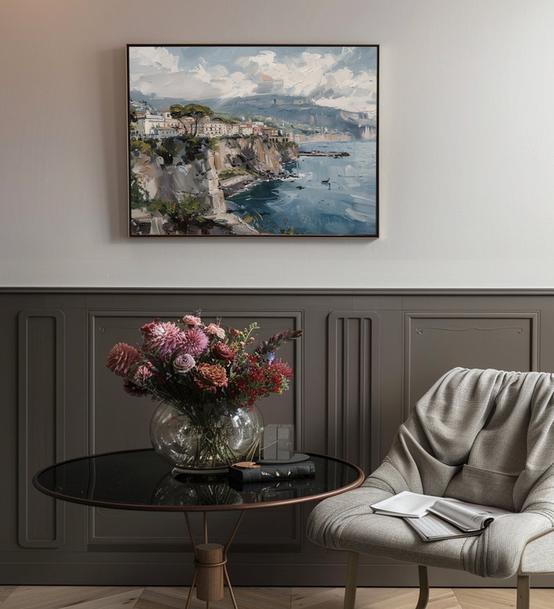 May include: A framed painting of a coastal town with buildings on a cliff overlooking a blue ocean. The painting is in a light wood frame and is hanging on a gray wall.