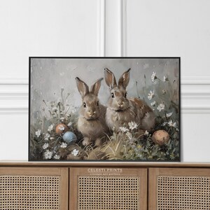 May include: Two brown rabbits sitting in a field of white flowers with blue and orange Easter eggs. The painting is in a black frame and has the text "Celesti Prints vintage art gallery" at the bottom.