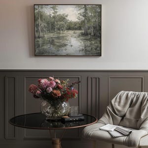 May include: A framed painting depicting a swamp scene with tall trees and still water. The painting is hung on a grey wall above a round table with a vase of pink and red flowers. A grey chair with a book on the seat is in the foreground.