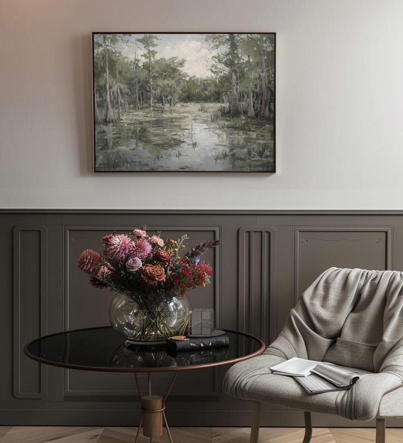 May include: A framed painting depicting a swamp scene with tall trees and still water. The painting is hung on a grey wall above a round table with a vase of pink and red flowers. A grey chair with a book on the seat is in the foreground.