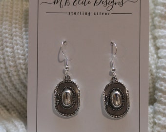 May include: A pair of silver earrings with a cowboy hat design. The earrings are on a white card with the text "MB Elite Designs - sterling silver".