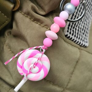 以下が含まれることがあります： ピンクと白の渦巻き型のロリポップチャームと、ピンクと白のビーズキーホルダー。キーホルダーはシルバーのキーリングに付いています。