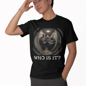 May include: Black t-shirt featuring a humorous image of three world leaders in a circular frame. The text "WHO IS IT?" is printed in white below the image. The shirt is made of a soft, dark fabric.