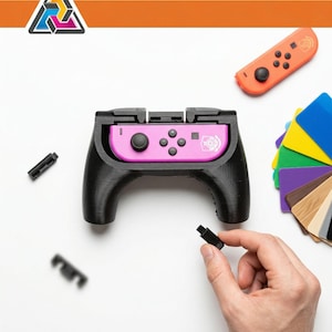 Puede incluir: Empuñadura de mando negra con centro rosa, diseñada para una consola de juegos. También se ven un mando naranja y muestras de color. Una mano sostiene una pequeña pieza negra. El logotipo está en la esquina superior izquierda.