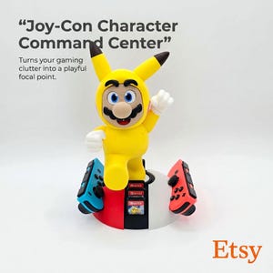May include: A Joy-Con Character Command Center stand featuring a yellow character figure wearing a hood with ears, standing on a red and white base. The base holds two game controllers and three game cartridges. Text reads Joy-Con Character Command Center.