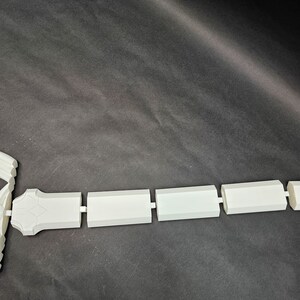 May include: A white, segmented object with a geometric design. The object is composed of rectangular and diamond-shaped components, connected in a linear fashion. The item is set against a dark background.