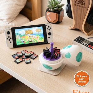 Op de afbeelding: Een witte Nintendo Switch-console met het spel "Animal Crossing" op het scherm. Naast de console staat een witte en turquoise spelcassettehouder met verschillende spelcassettes. Een houten hoofdtelefoonstandaard en een gepixeleerde plantenpot zijn ook zichtbaar.