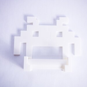 May include: White plastic toilet paper holder shaped like a pixelated space invader.
