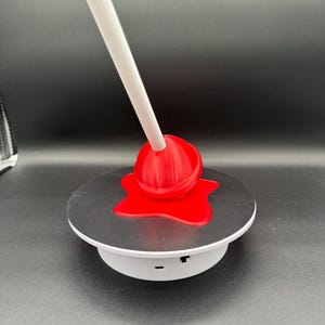 May include: A red lollipop with a white stick, resting on a black and white circular platform. The lollipop appears to be melting, with a red, stylized puddle beneath it. The platform is on a black background.