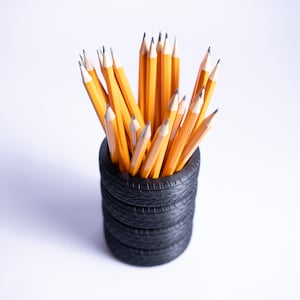 Może przedstawiać: Czarny, oponowy pojemnik na ołówki wypełniony żółtymi ołówkami.