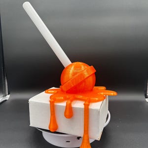 May include: An orange 3D-printed lollipop design with a white stick, appearing to drip over a white box. The lollipop is a sphere with a ring around it. The dripping effect is a stylized, melted design.