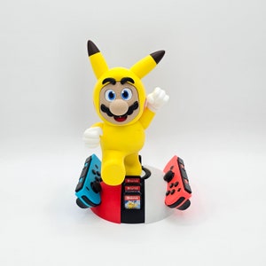 Può includere: Una statuetta di Mario a tema Pikachu gialla si trova su una base bianca e blu con due controller Nintendo Switch rossi e blu. La base ha uno slot per tre cartucce di gioco.