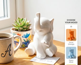 May include: A white ceramic elephant figurine with its trunk raised, sitting on a wooden surface. A small card with the words "for good luck" is in front of it. The image also includes a mug, a potted succulent, and a color chart with the text "CHOOSE YOUR MOOD".