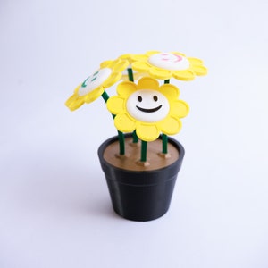 May include: A decorative potted plant featuring yellow flowers with smiling faces. The flowers have white centers and green stems, set in a black pot. The pot is filled with a brown material, and the background is plain white.