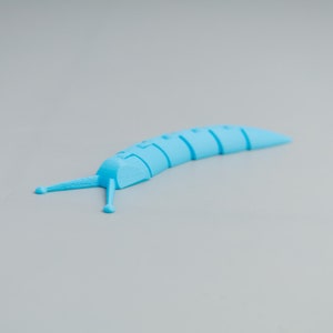 Könnte beinhalten: Ein hellblaues, segmentiertes, 3D-gedrucktes Schneckenspielzeug. Das Spielzeug hat einen gebogenen Körper mit einer Reihe von verbundenen Segmenten, zwei Antennen und einem spitzen Schwanz. Das Spielzeug liegt auf einer grauen Oberfläche.