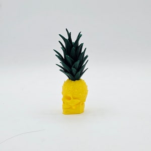 May include: A yellow pineapple skull with a green leafy top. The skull has detailed facial features and a textured surface. The pineapple skull is a unique decorative item.