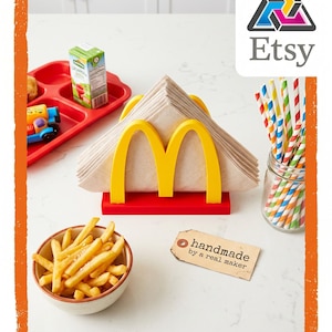 May include: A red and yellow napkin holder with the McDonald's logo, holding folded napkins. A bowl of french fries, a red tray with a juice box and toy, and a jar of colorful straws are also present. A tag reads "handmade by a real maker."