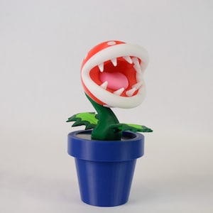May include: A red and white plant with a large open mouth and sharp teeth, in a blue pot. The plant is a popular character from the Super Mario video game series.