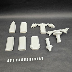 May include: A collection of white 3D-printed components, including blade-like shapes, rectangular pieces, and a handle-like structure. The parts are arranged on a dark surface, suggesting a model kit or prototype.