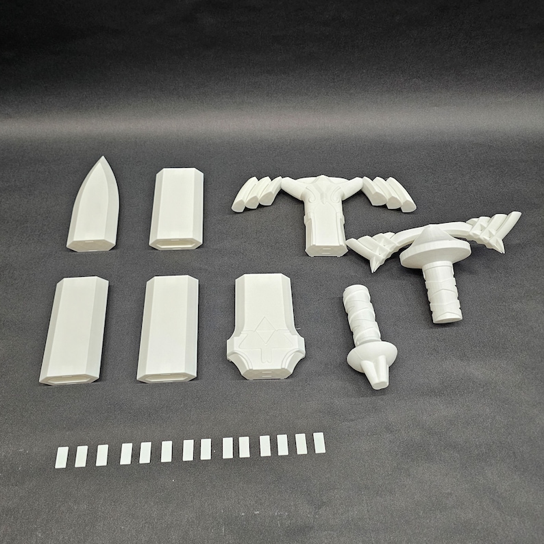 May include: A collection of white 3D-printed components, including blade-like shapes, rectangular pieces, and a handle-like structure. The parts are arranged on a dark surface, suggesting a model kit or prototype.