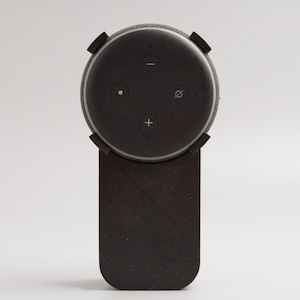Puede incluir: Un soporte de pared negro para un altavoz inteligente con un altavoz circular adjunto. El altavoz tiene cuatro botones: un botón de micrófono, un botón para subir el volumen, un botón para bajar el volumen y un botón con un círculo en su interior.