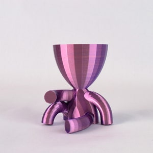 Könnte beinhalten: Eine violette und rosafarbene gestreifte Keramikvase mit abgerundeter Oberseite und drei geschwungenen Beinen. Die Vase ist aus einem glatten, strukturierten Material gefertigt.