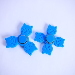 Puede incluir: Dos spinners azules con diseño de cara de gato. Cada spinner tiene tres brazos con forma de gato y un botón central azul. Los spinners son de plástico y están diseñados para girar y jugar.