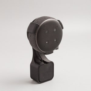 Puede incluir: Un altavoz negro con una cubierta de tela y un soporte negro. El altavoz tiene un micrófono y controles de volumen. El soporte tiene una base cuadrada y un brazo curvo.