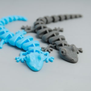 Peut inclure: Deux lézards en plastique, l'un bleu et l'autre gris, sont posés sur une surface grise. Les lézards sont segmentés et ont une apparence squelettique.