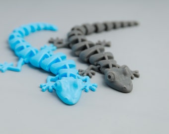 May include: Two plastic lizards, one blue and one gray, are posed on a gray surface. The lizards are segmented and have a skeletal appearance.