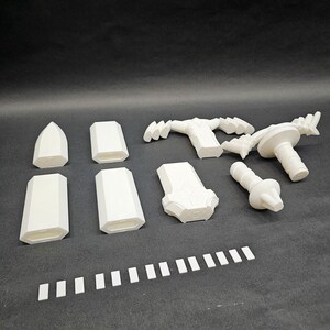 May include: A collection of white 3D-printed components, including geometric shapes, handles, and a cylindrical piece with a knob. The parts are arranged on a dark surface, suggesting a kit or set of modular elements.